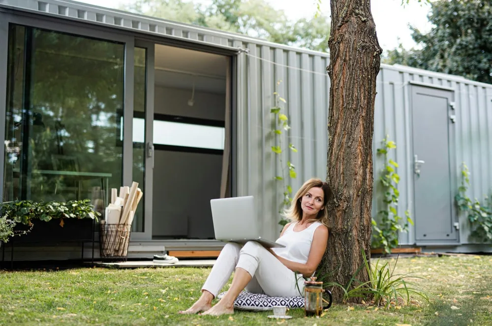 Femme assise dans un jardin avec un ordinateur portable, devant un bâtiment container doté d'une large ouverture vitrée.